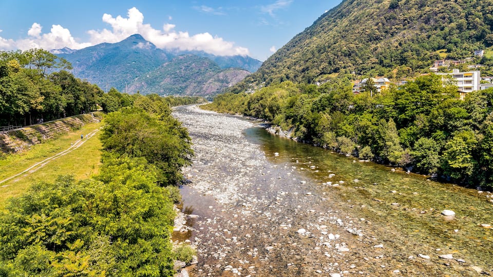 A river flowing through a mountainous area with dense greenery and a few buildings on the right.