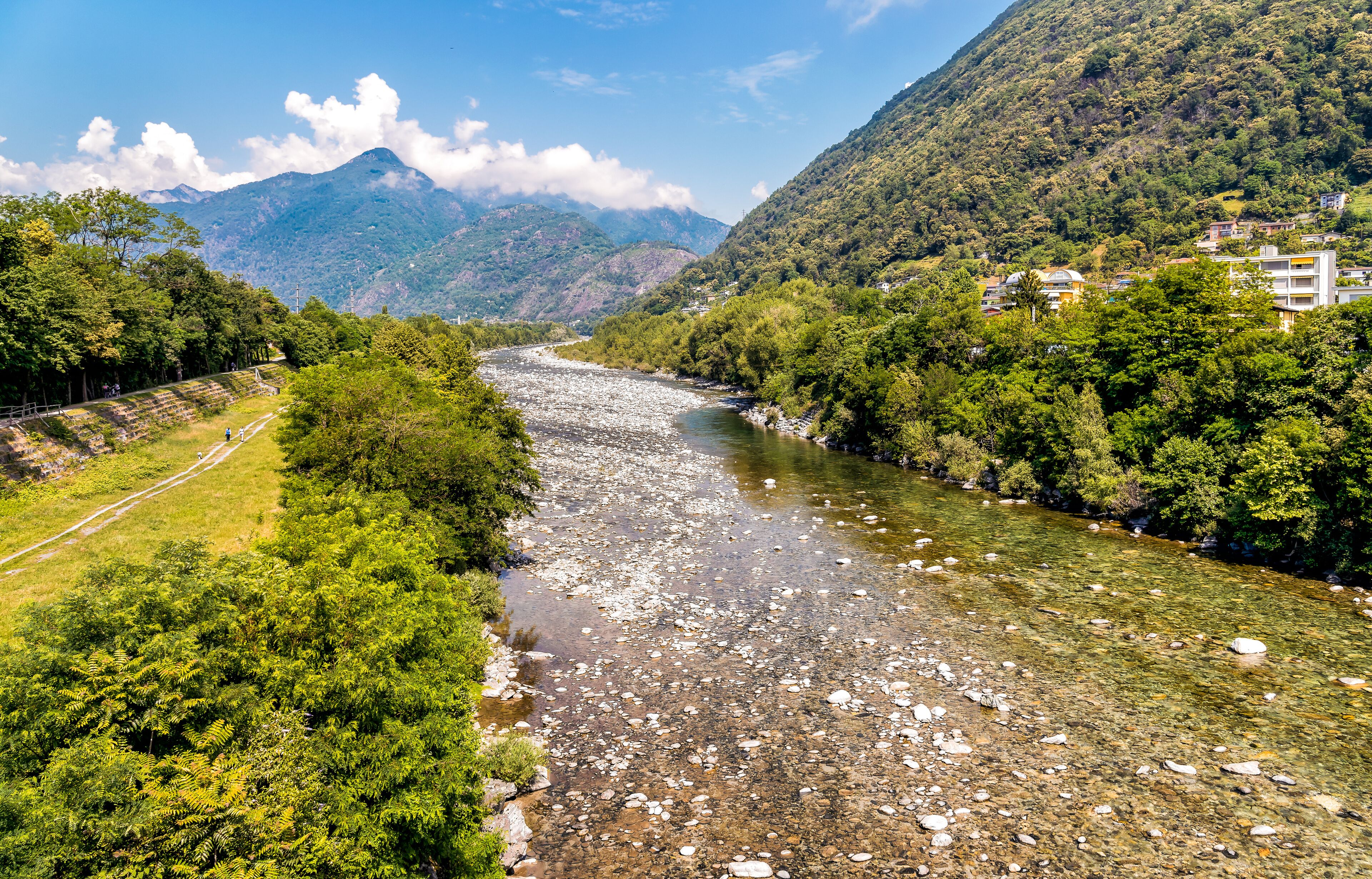 A river flowing through a mountainous area with dense greenery and a few buildings on the right.