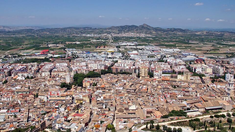 A densely built urban area with a mix of historical and modern architecture, surrounded by green fields and distant mountains.