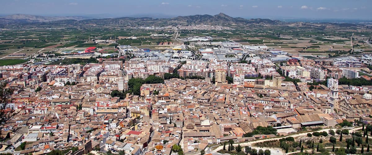 A densely built urban area with a mix of historical and modern architecture, surrounded by green fields and distant mountains.