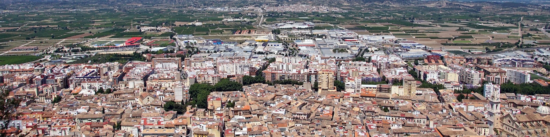 A densely built urban area with a mix of historical and modern architecture, surrounded by green fields and distant mountains.