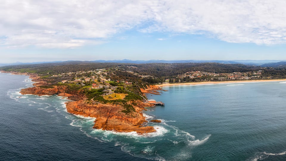 An aerial view of a coastal town with a beach, rocky coastline, and surrounding hills.