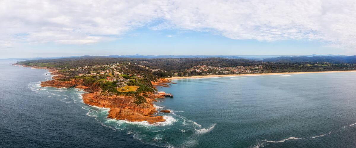An aerial view of a coastal town with a beach, rocky coastline, and surrounding hills.