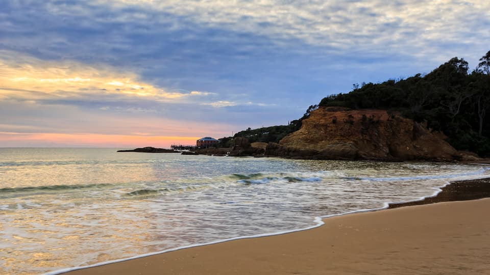 A beach with waves, a rocky cliff, and a sunset.