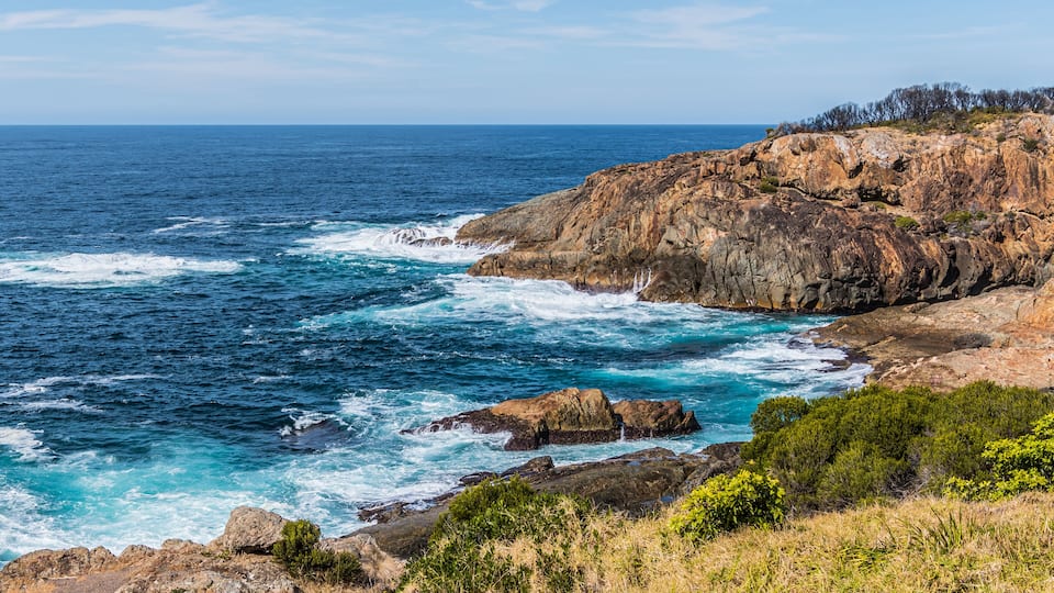 A coastal landscape with rocky cliffs, ocean waves, and sparse vegetation.