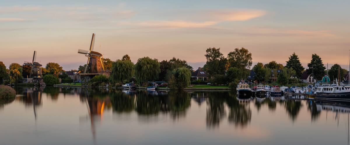 A calm river with a windmill, boats, and trees reflecting in the water.
