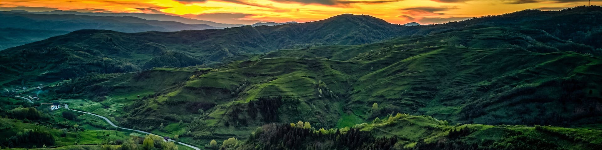 A winding road through a lush green valley with rolling hills and a sunset in the background.