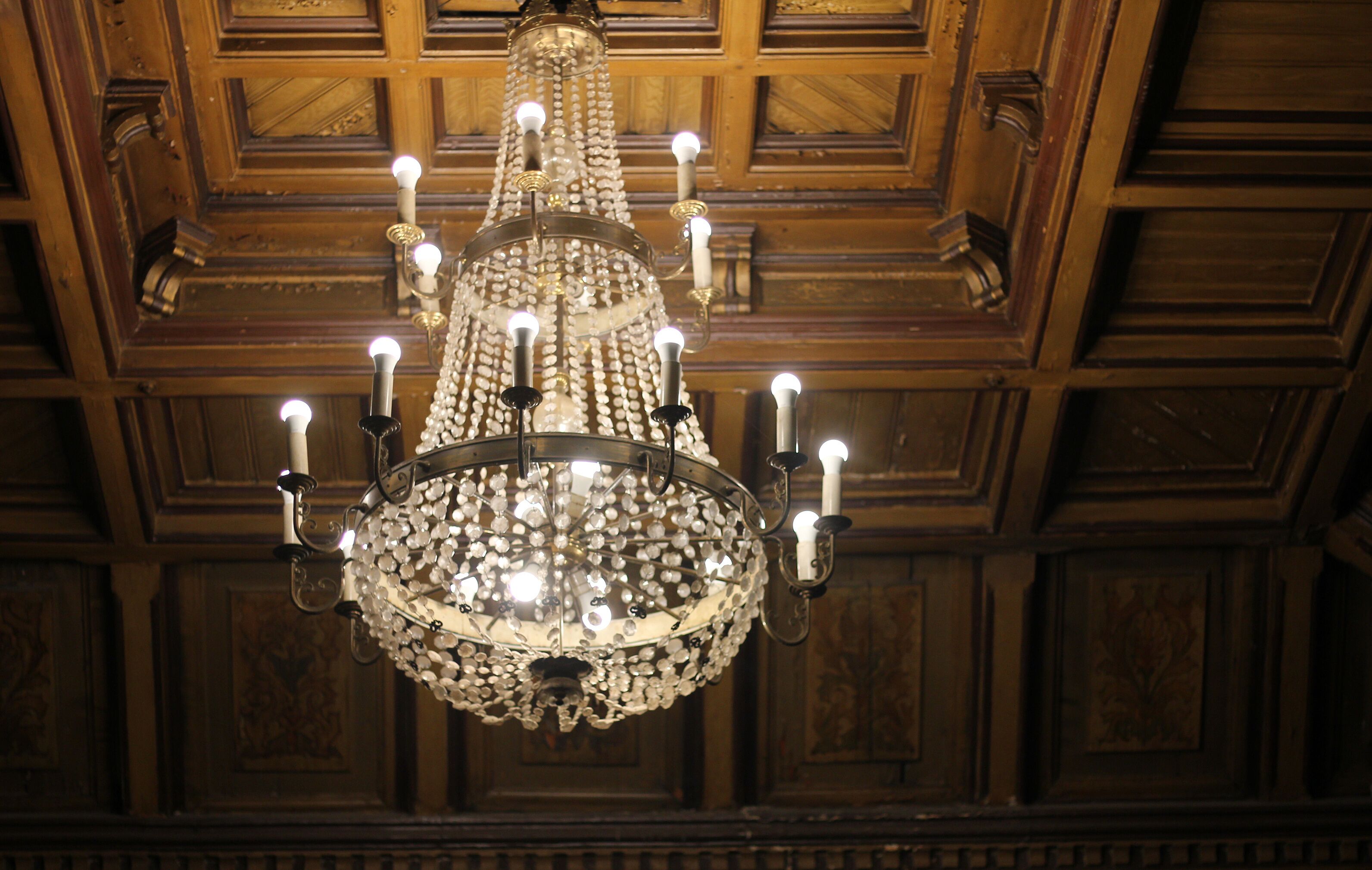 Decorated chandelier hangs in the entrance hall at Odessa Philharmonic. Historical large three level brass chandelier with 116 arms on the background of decorated wooden ceiling. 16th century artwork