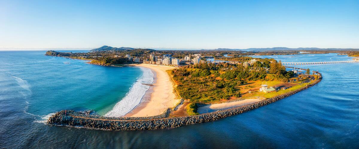 A coastal cityscape with a beach, a bridge, and a peninsula with buildings and greenery.