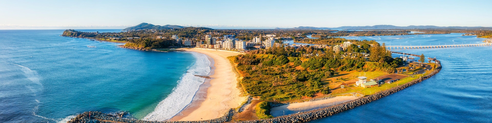 A coastal cityscape with a beach, a bridge, and a peninsula with buildings and greenery.