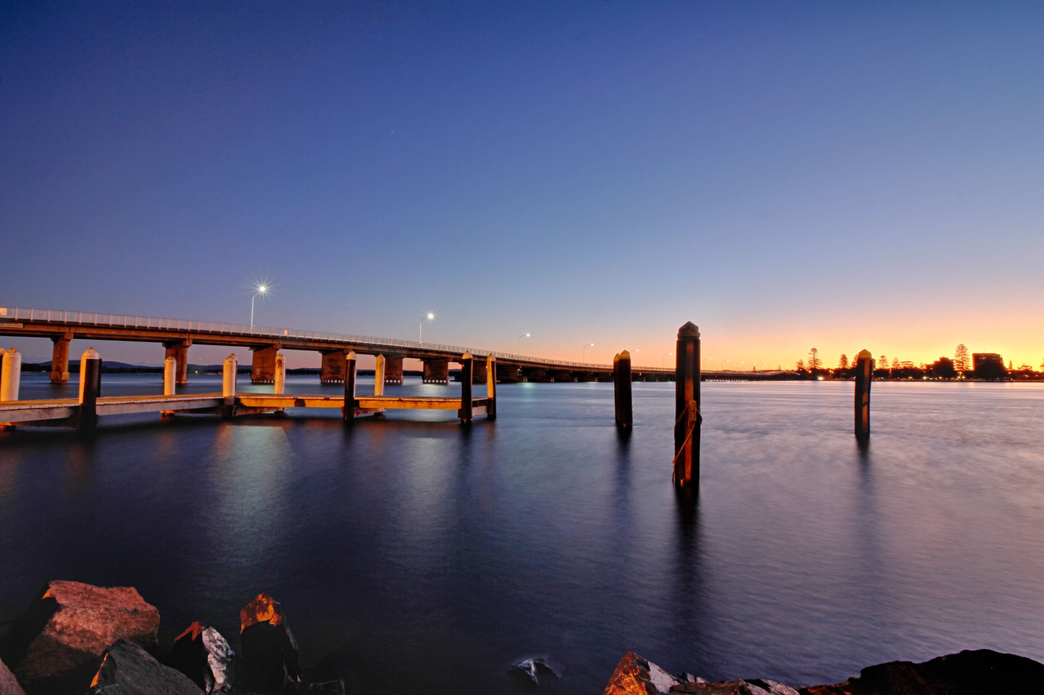 A bridge over calm water at dusk with streetlights and a sunset in the background.