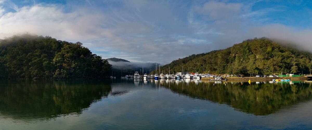 Beautiful morning panoramic view of Cockle creek with reflections of blue sky, light clouds, foggy mountains and trees, Bobbin Head, Ku-ring-gai Chase National Park, New South Wales, Australia