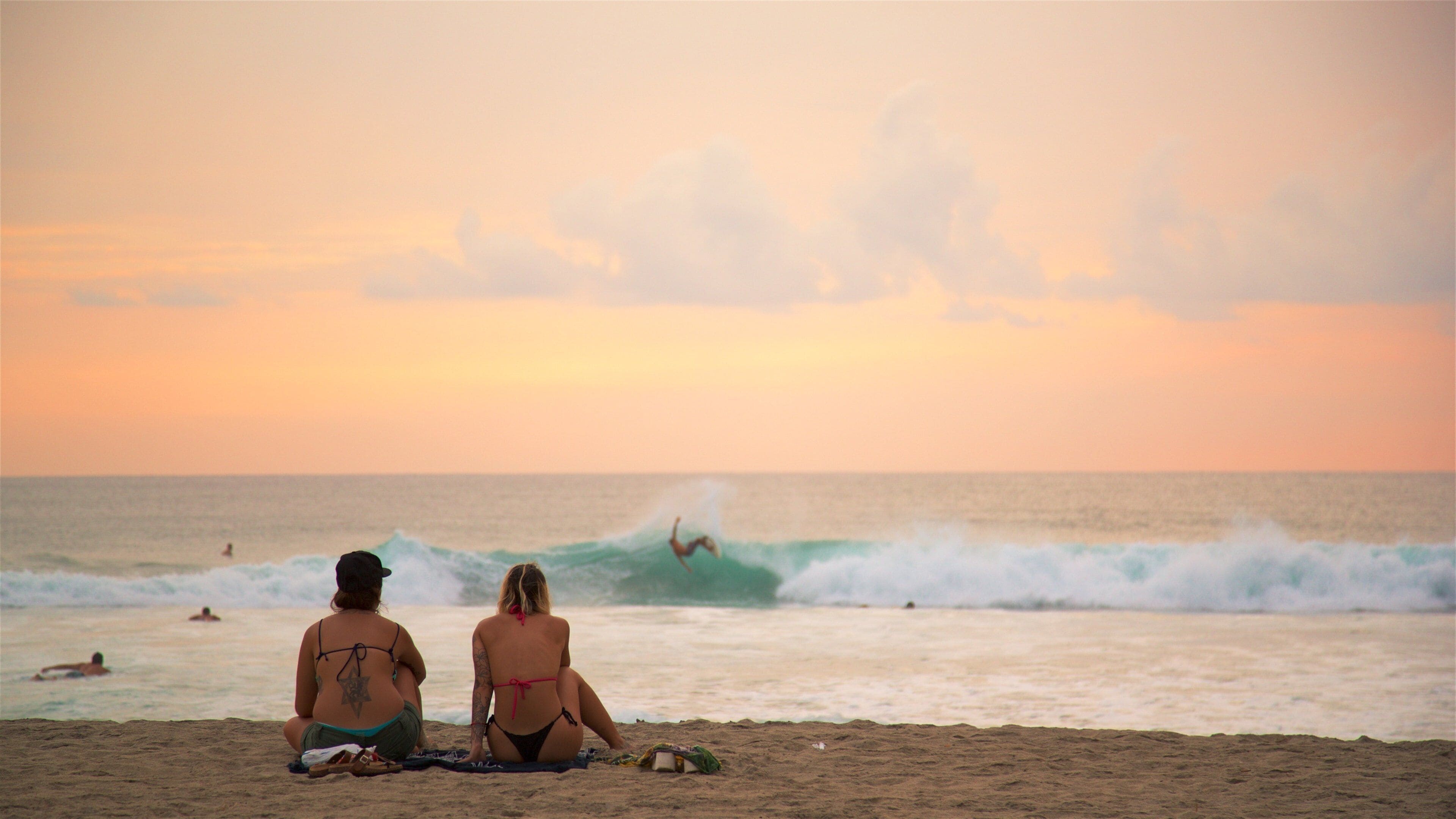 Zwei Personen sitzen am Strand und beobachten einen Surfer im Meer bei Sonnenuntergang.