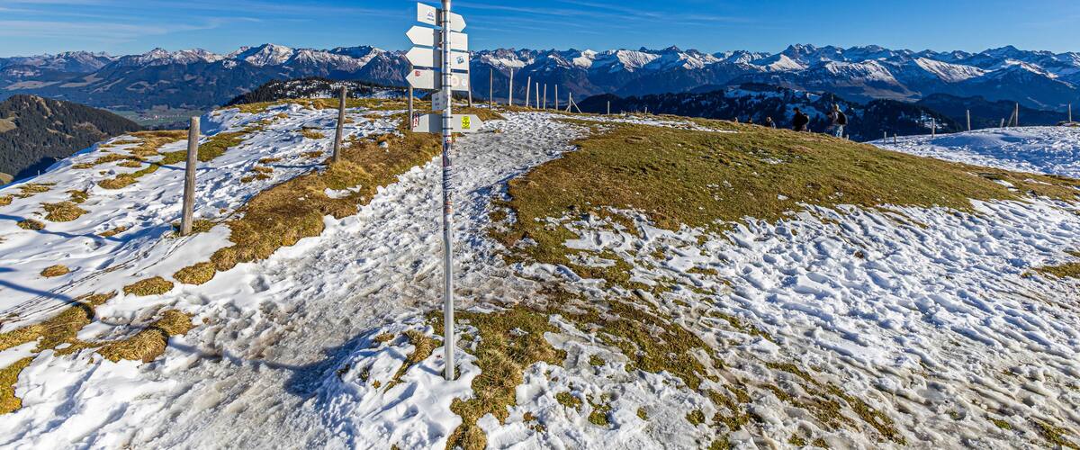 A snow-covered mountain landscape with a signpost and distant snow-capped peaks under a clear blue sky.