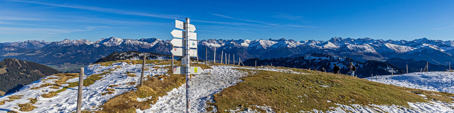 A snow-covered mountain landscape with a signpost and distant snow-capped peaks under a clear blue sky.