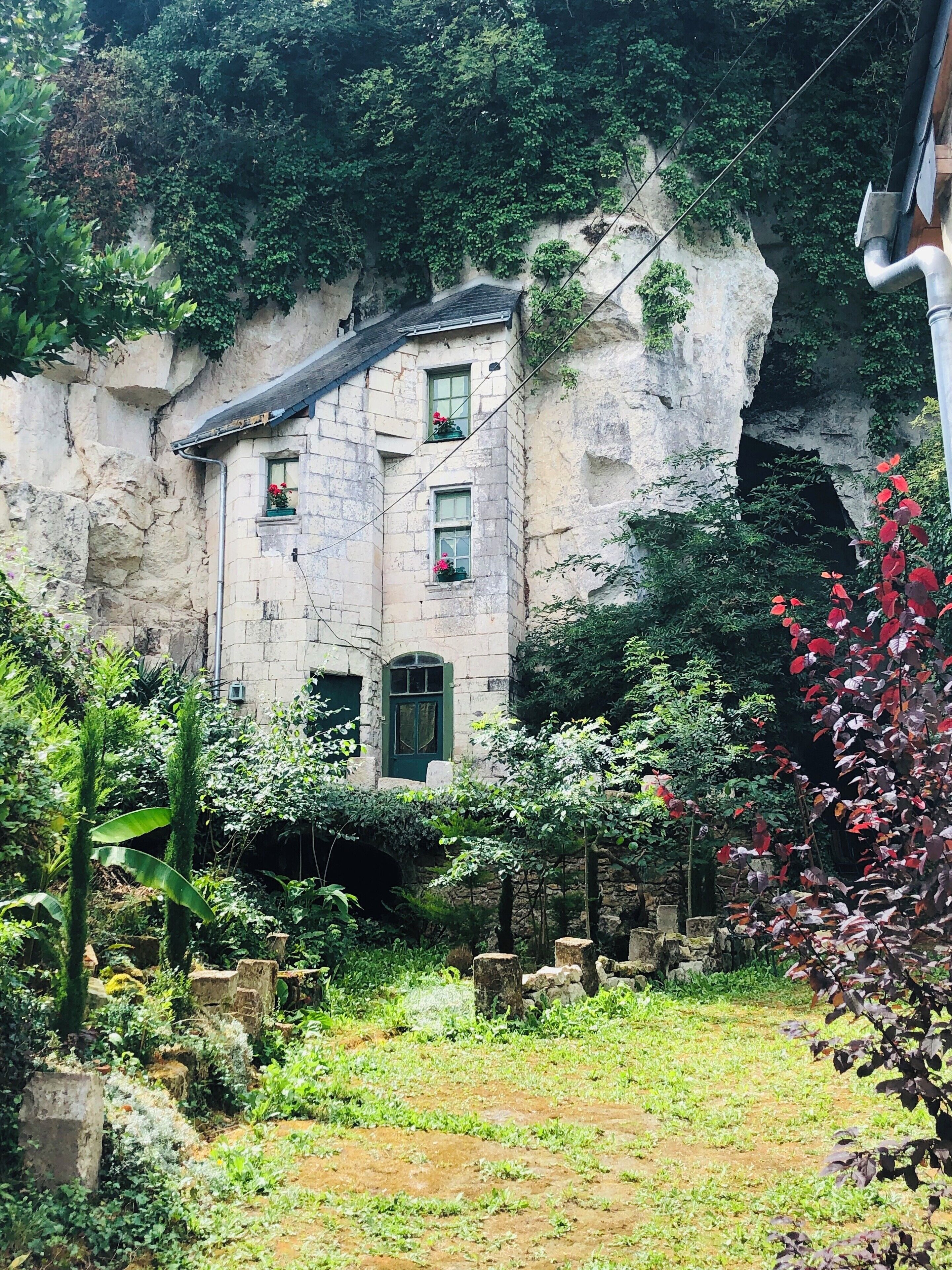 A stone building with windows and a door, surrounded by lush greenery and a rocky cliff.