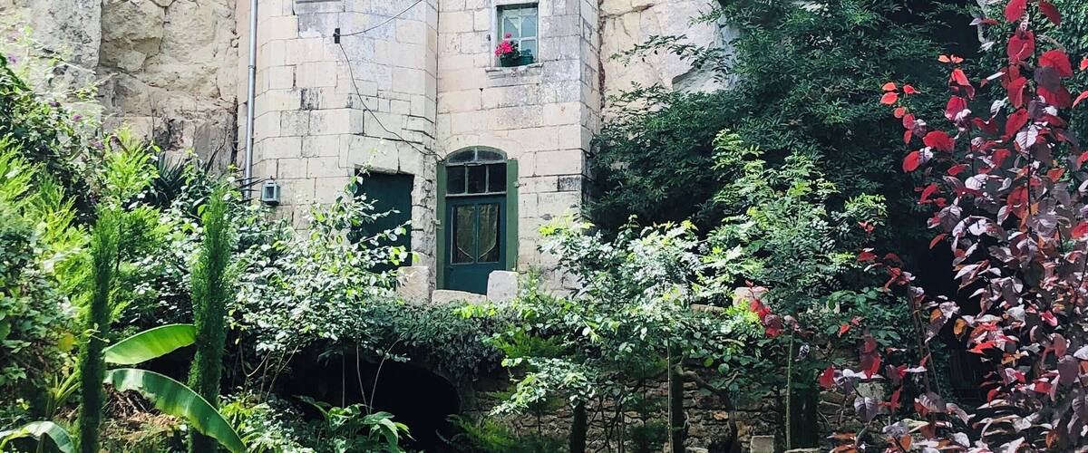 A stone building with windows and a door, surrounded by lush greenery and a rocky cliff.