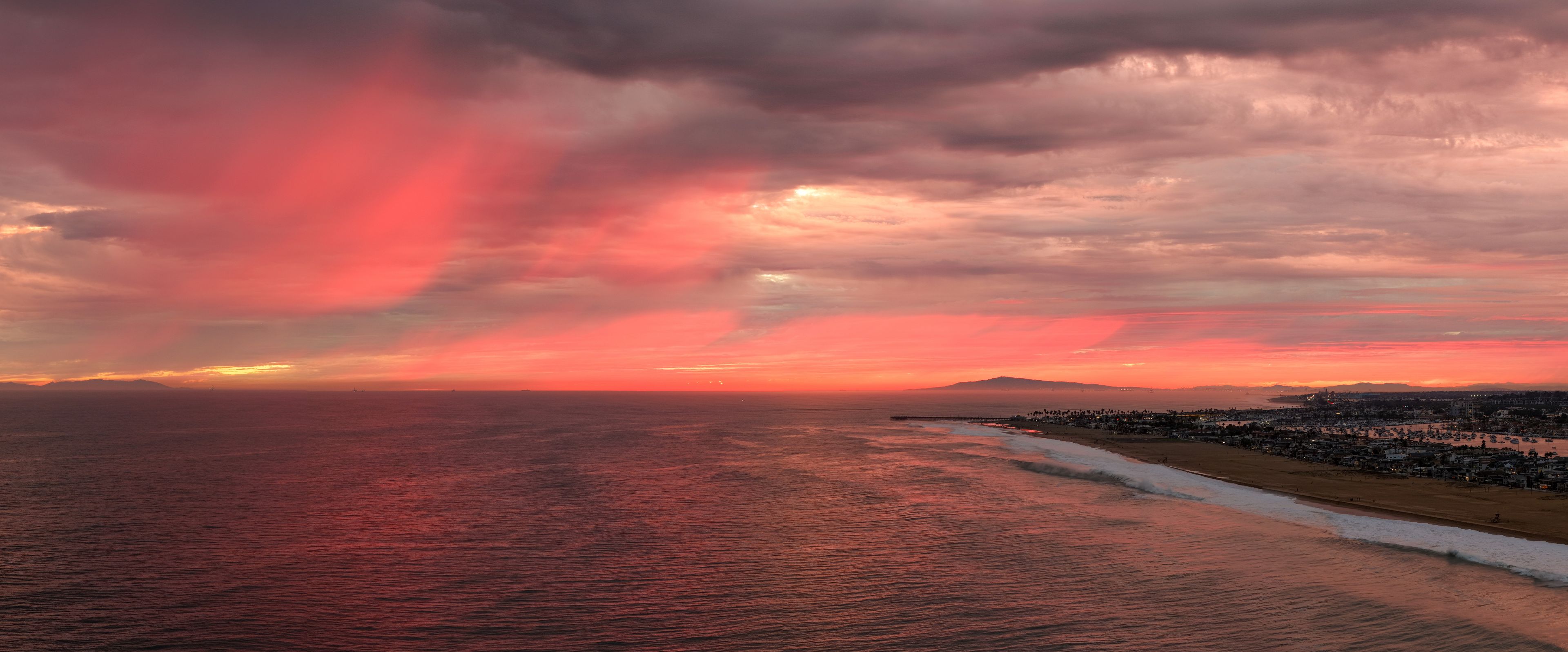 Dramatic red sunset beneath dark storm clouds over the Pacific Ocean and Newport Beach coastline in Southern California, with glowing crimson light stretching across the horizon and reflecting on the 
