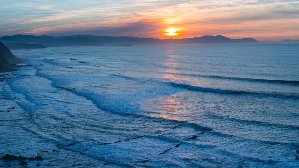 A coastal sunset with waves, cliffs, and distant mountains.