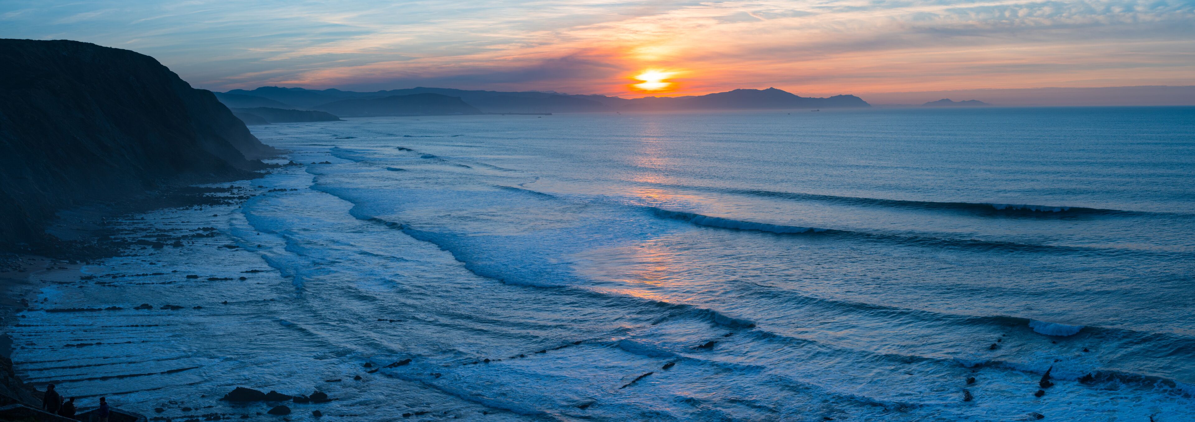 A coastal sunset with waves, cliffs, and distant mountains.