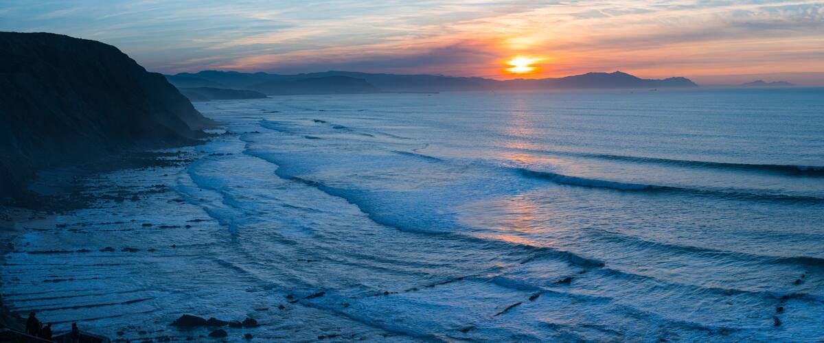 A coastal sunset with waves, cliffs, and distant mountains.