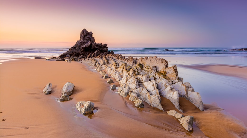 A beach with a rock formation and waves at sunset.