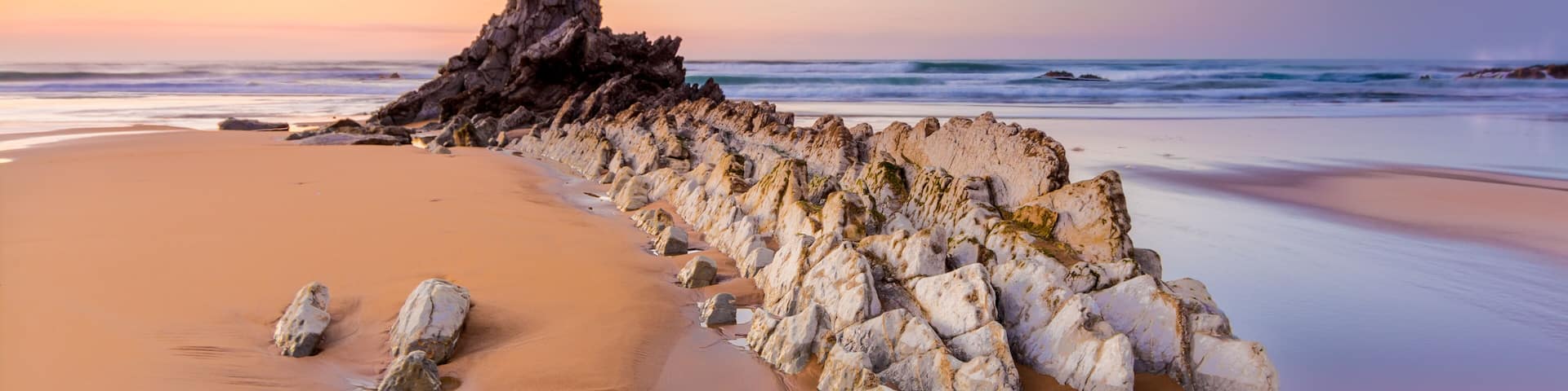A beach with a rock formation and waves at sunset.