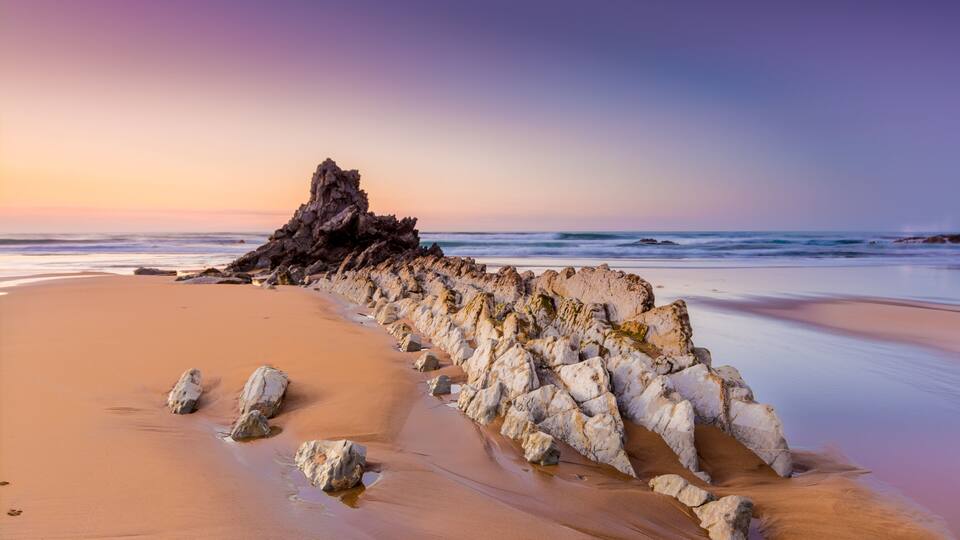 A beach with a rock formation and waves at sunset.