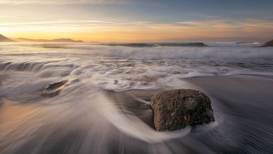 A rocky shoreline with waves crashing onto the beach during sunset.