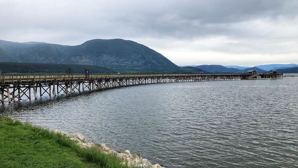 A wooden pier extending into a calm lake, surrounded by grassy banks and mountains in the background.