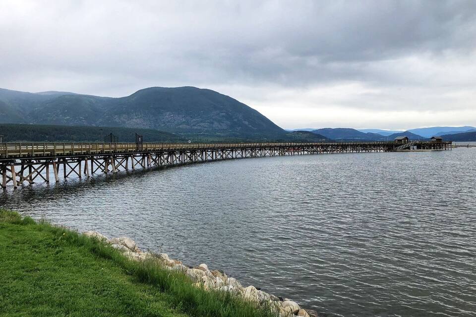 A wooden pier extending into a calm lake, surrounded by grassy banks and mountains in the background.