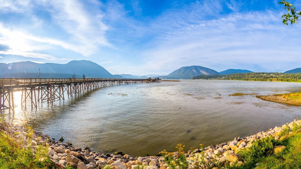 A river with a wooden bridge, a grassy bank, and mountains in the background.