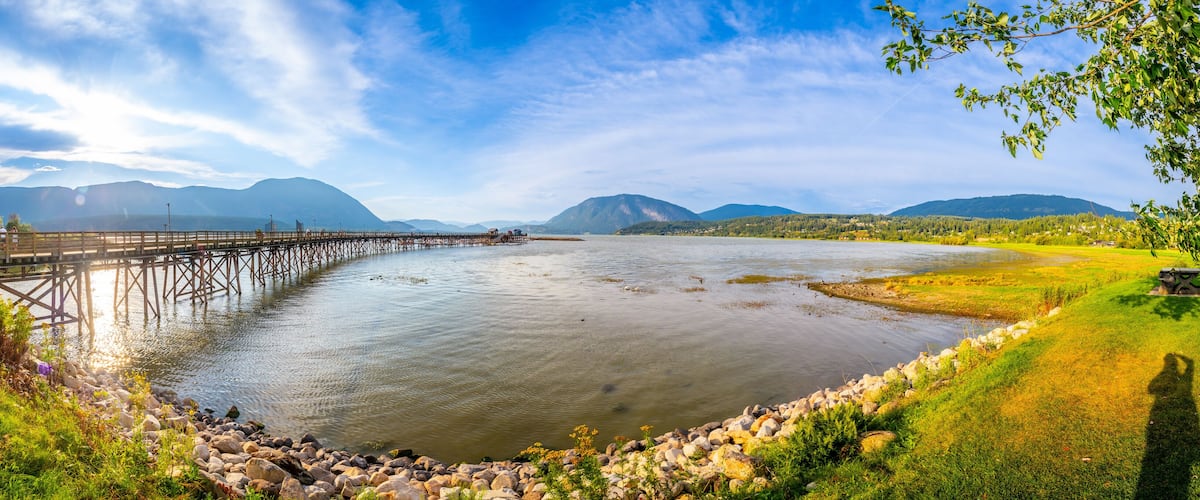 A river with a wooden bridge, a grassy bank, and mountains in the background.