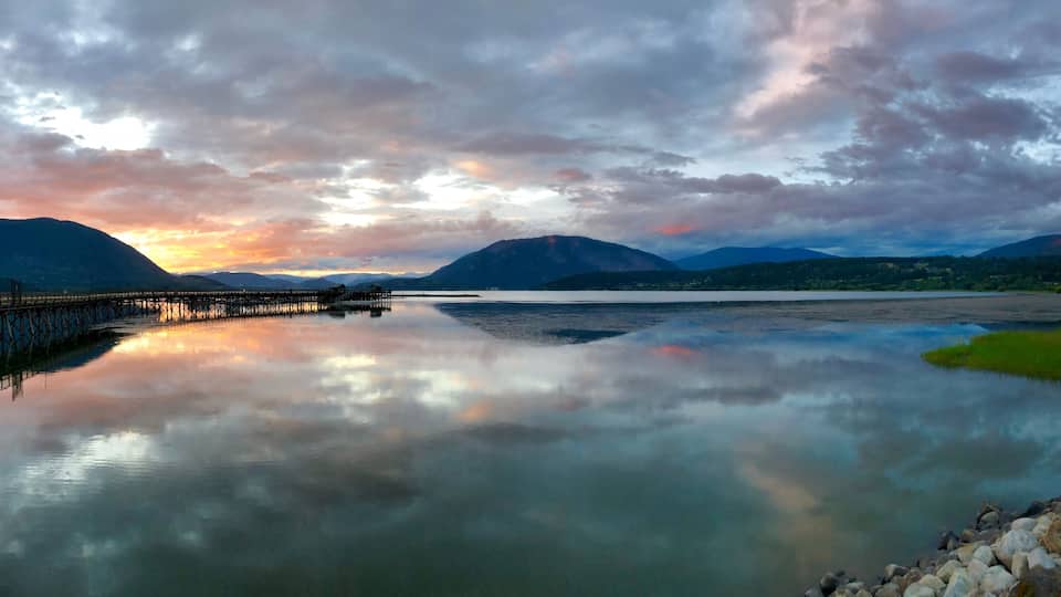 A calm lake with a pier extending into it, surrounded by mountains and a cloudy sky.