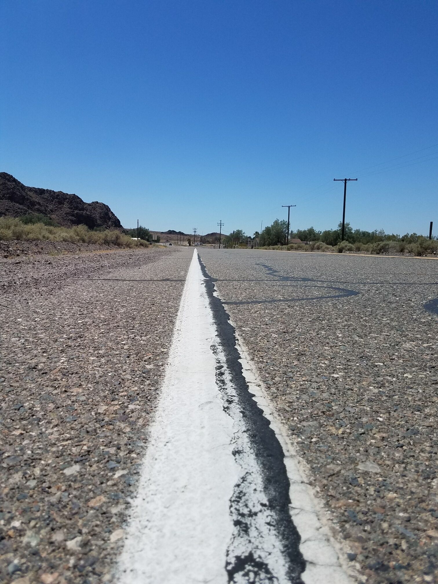 A gravel road with a white line in the center, surrounded by sparse vegetation and hills in the distance.