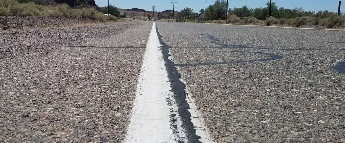 A gravel road with a white line in the center, surrounded by sparse vegetation and hills in the distance.