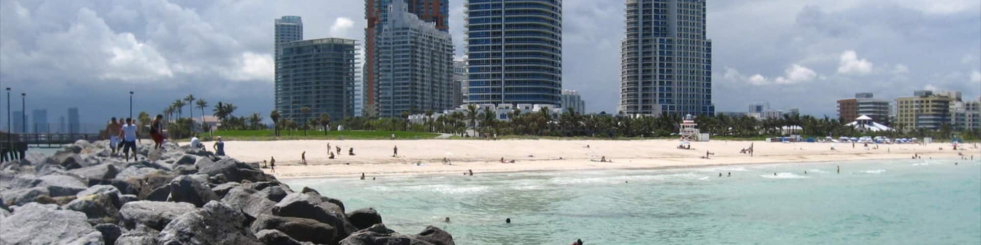 A beach with clear turquoise water, a rocky shoreline, and several high-rise buildings in the background.