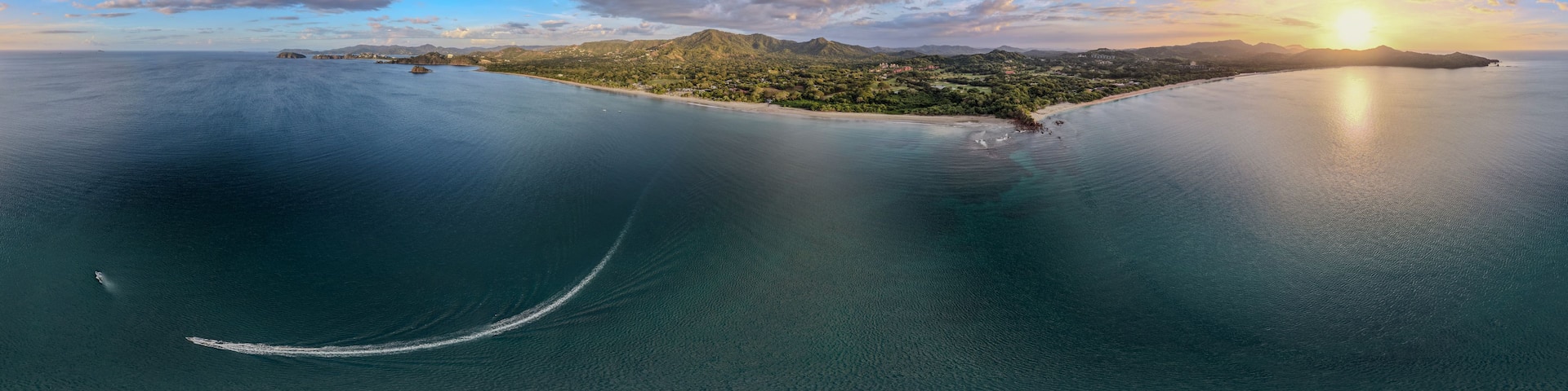 A coastal landscape with a clear sky, a calm sea, and a sun setting over a mountainous island.