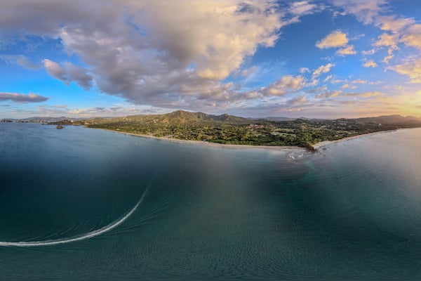 Paisagem costeira com céu claro, mar calmo e sol se pondo sobre uma ilha montanhosa.