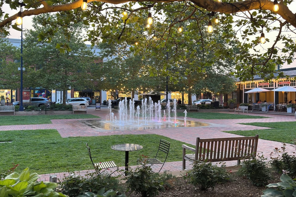 Una fontana in un parco con panchine e tavoli, circondata da alberi e illuminata da luci a catena.