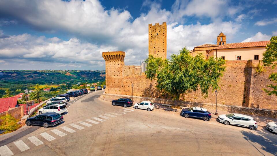 A historic castle with a tall tower, a parking lot with cars, and a scenic view of the surrounding hills.