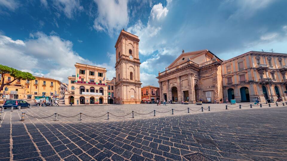 A historic square with a prominent clock tower, cobblestone pavement, and classical architecture.