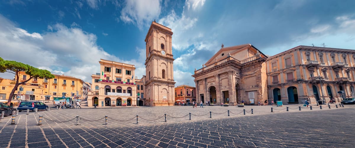 A historic square with a prominent clock tower, cobblestone pavement, and classical architecture.