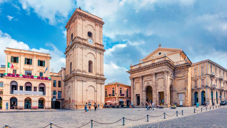 A historic square with a prominent clock tower, a large archway, and classical architecture.