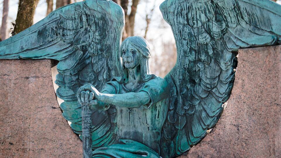 A bronze angel statue with wings, holding a staff, and seated on a stone base.