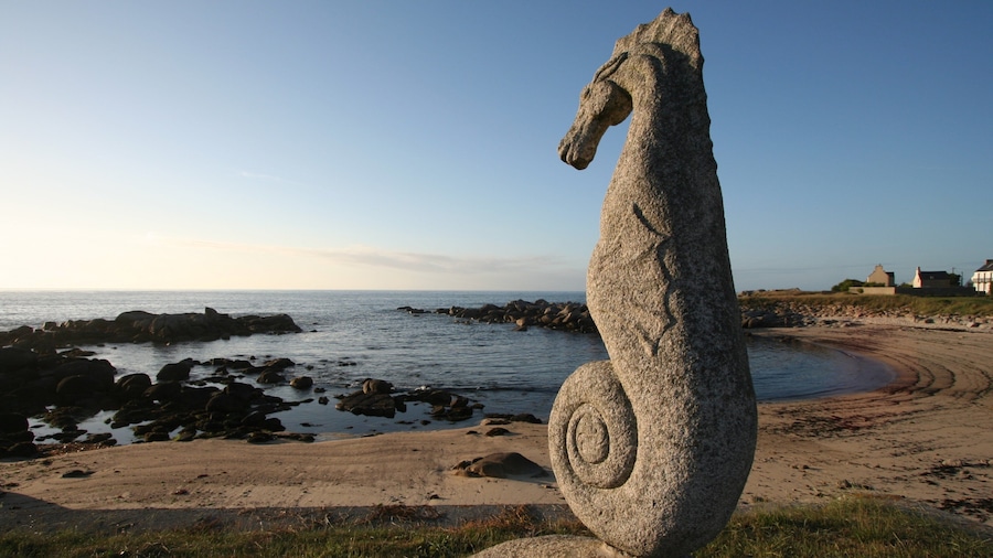 Una escultura de caballito de mar de piedra sobre una playa con costa rocosa y casas al fondo.