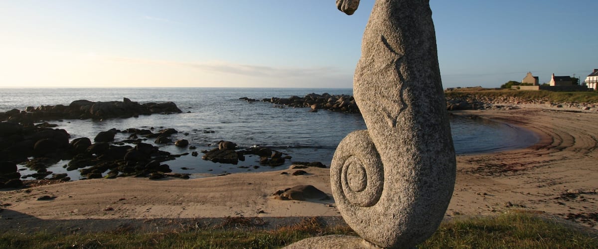 A stone seahorse sculpture on a beach with a rocky shoreline and houses in the background.