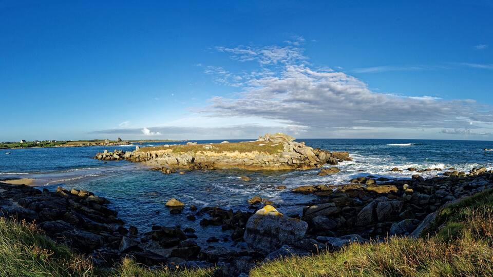 A rocky coastline with a prominent rock formation in the center, surrounded by ocean waves and a clear blue sky.