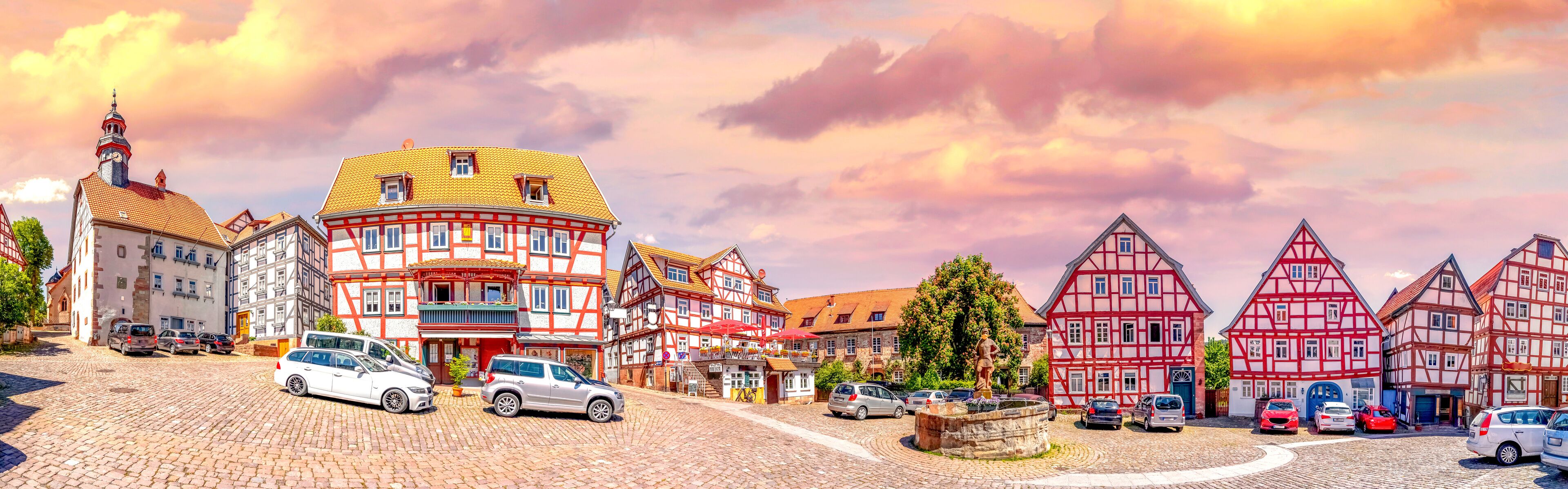 A cobblestone street lined with traditional half-timbered houses, a statue, and parked cars.