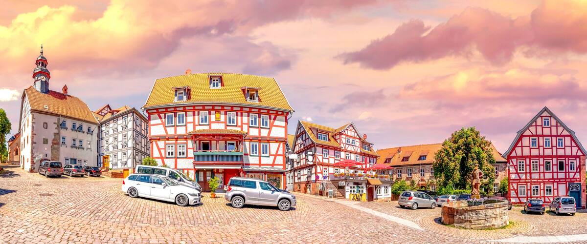 A cobblestone street lined with traditional half-timbered houses, a statue, and parked cars.